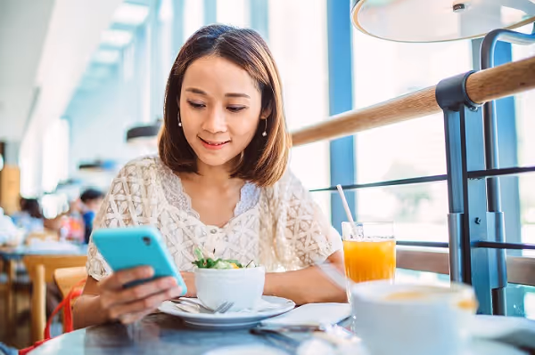 Pretty young Asian woman using smartphone joyfully while having meal in a restaurant.