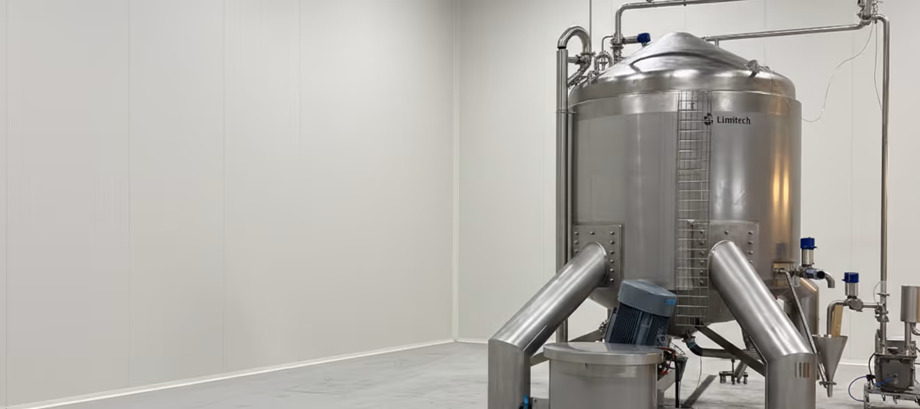 Large stainless steel industrial tank with pipes and machinery in a clean, white industrial room.