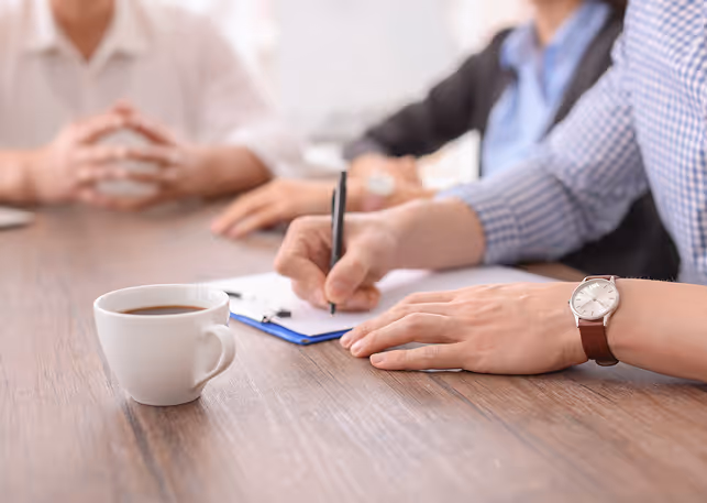 Person writing on clipboard at a table with teammates and a cup of coffee nearby.