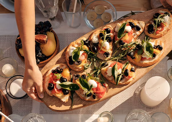 Hand holding a wooden tray with toasted bread topped with tomato, olives, cheese, and herbs.