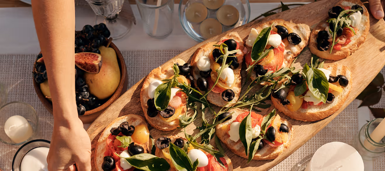 Hand serving bruschetta topped with tomatoes, mozzarella, olives, and basil on a wooden board.