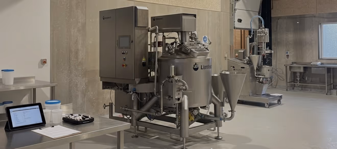 Industrial stainless steel mixer machine in a clean room with tables holding containers and a tablet.