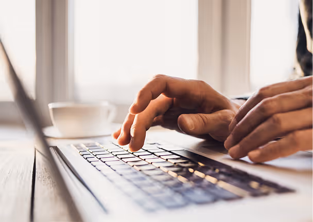 Hands typing on a laptop keyboard next to a white coffee cup by a window.