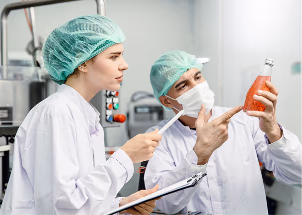 Two lab technicians in protective gear inspecting a bottle of red liquid in a lab or factory.
