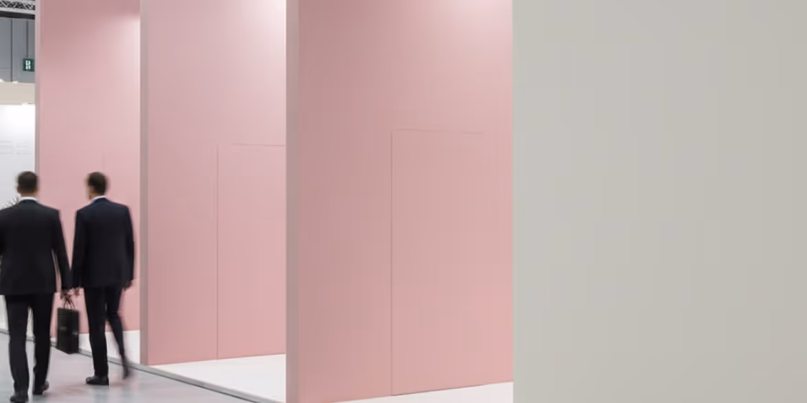 Two men in business suits walking past large pink partition walls in a modern indoor space.