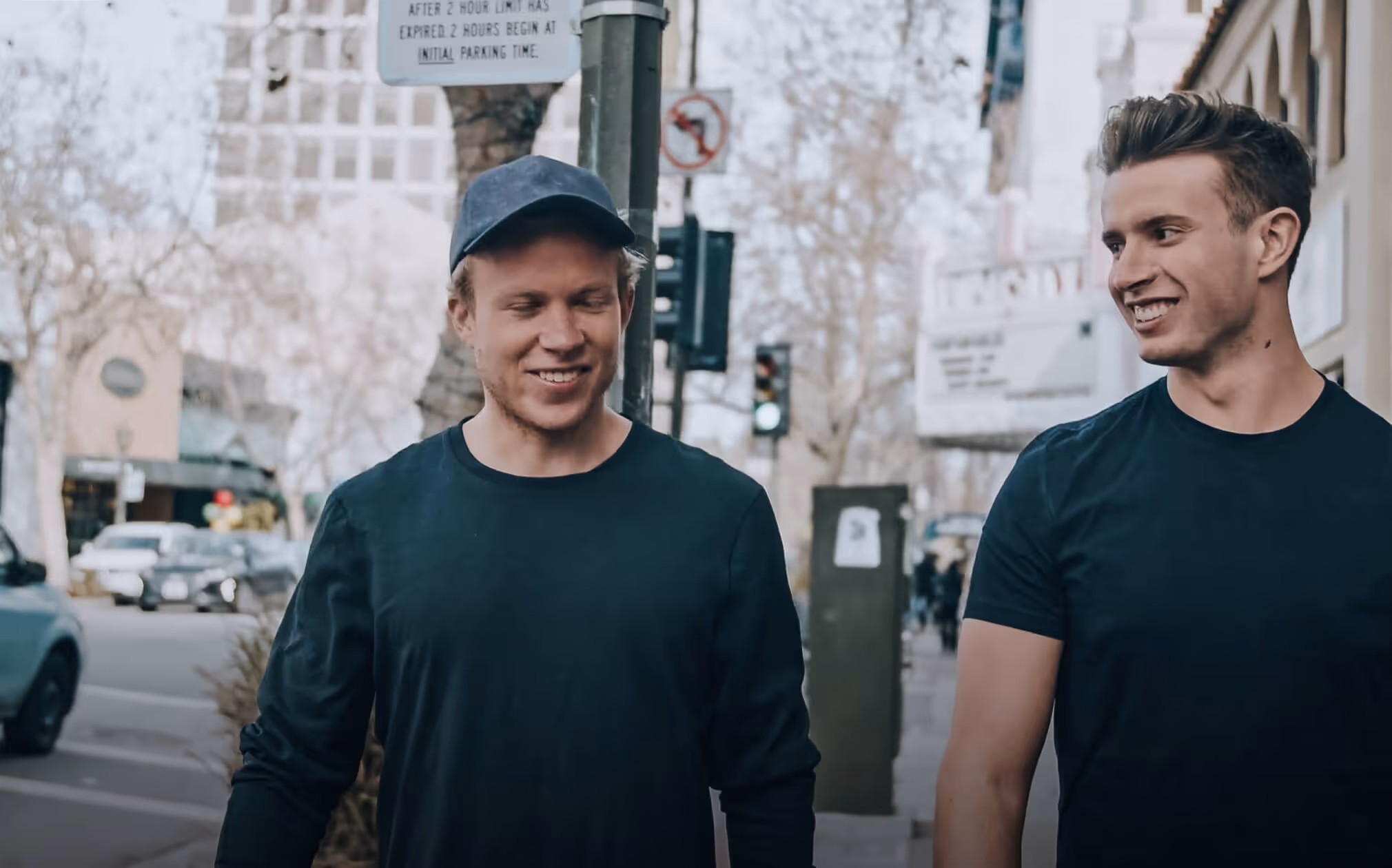 Two young men smiling and walking together on a city sidewalk with trees and buildings in the background.