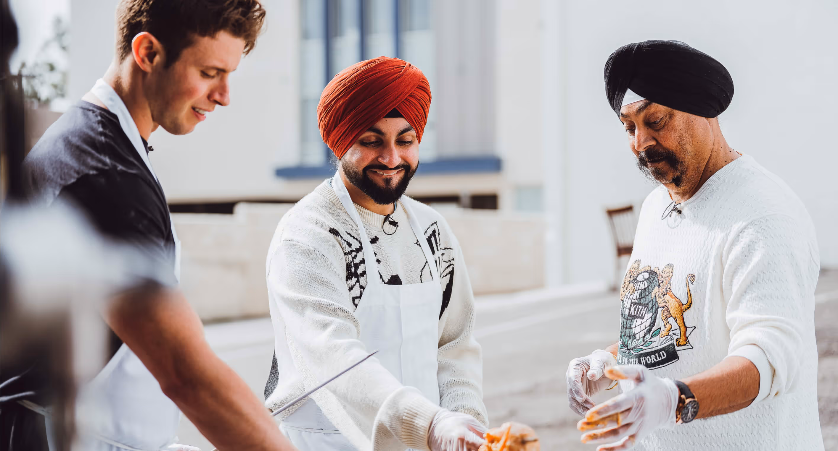 Three men, two wearing turbans and aprons, preparing food outdoors while smiling and engaging.