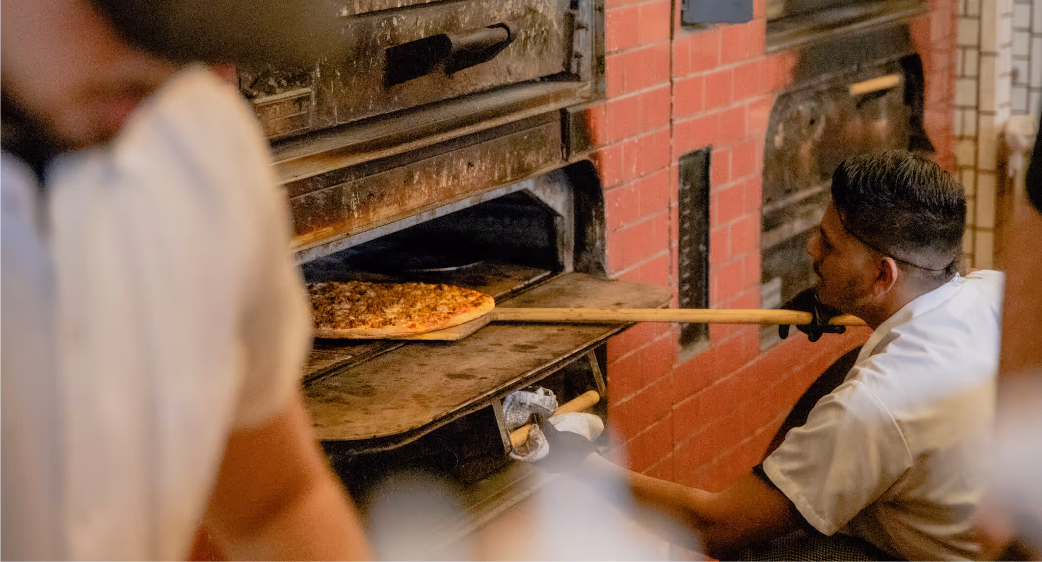 Chef using a wooden peel to place a pizza into a brick oven.