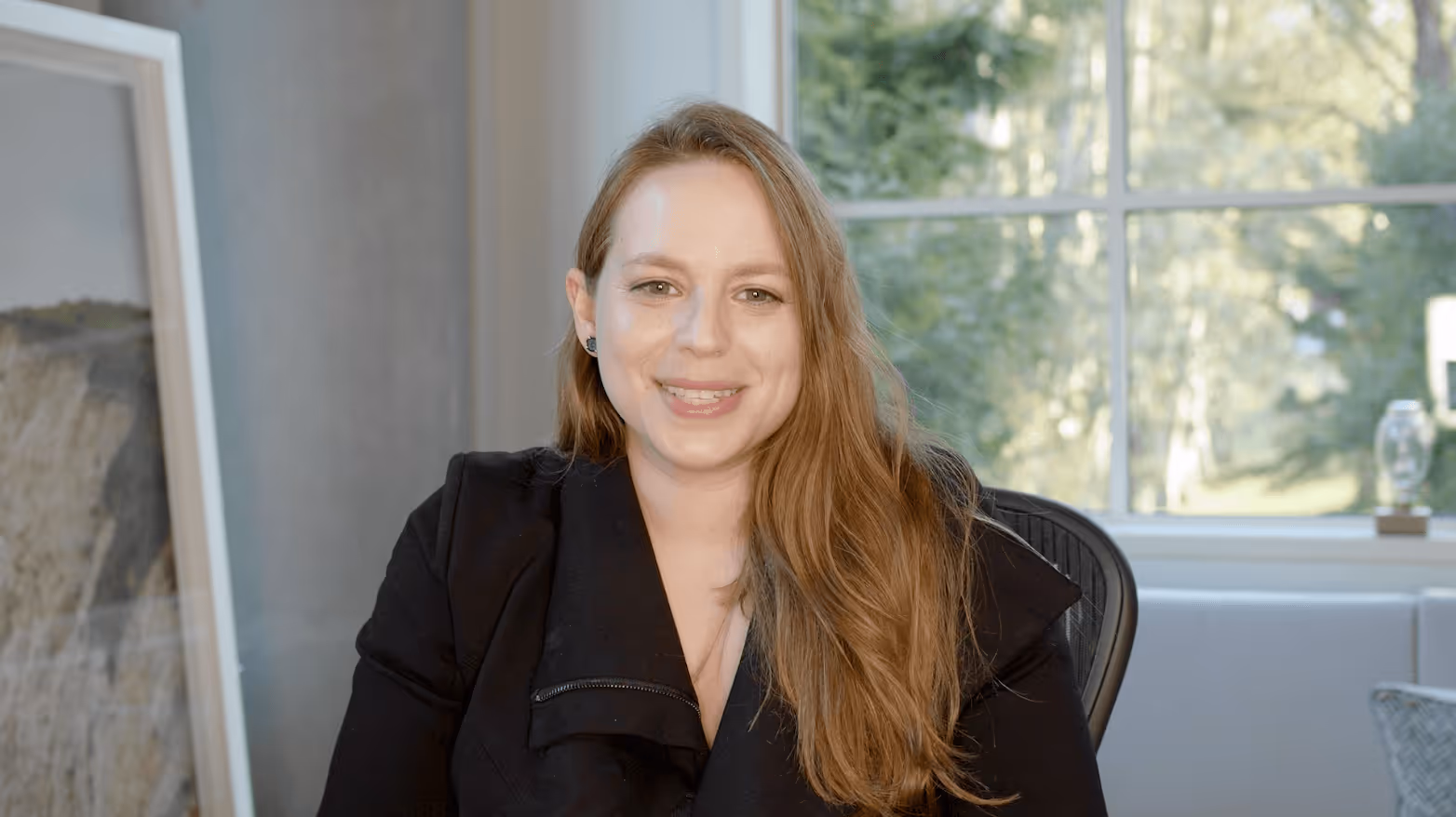 Smiling woman with long light brown hair wearing a black jacket sitting indoors with a window and greenery in the background.