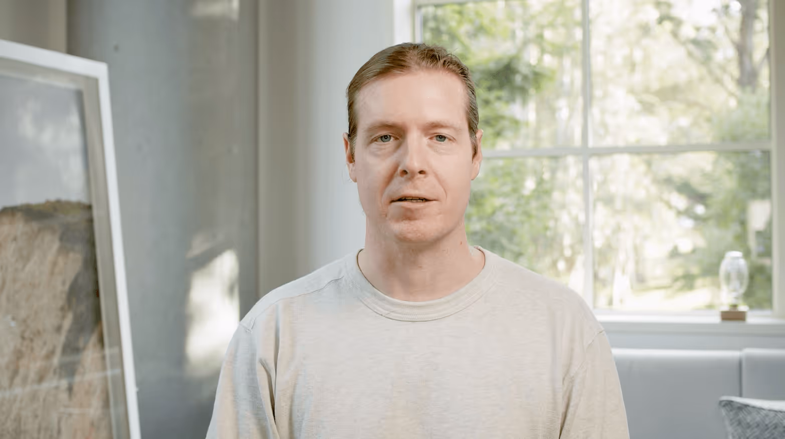 Man with light brown hair tied back wearing a light gray sweatshirt standing indoors in front of a large window with greenery outside.