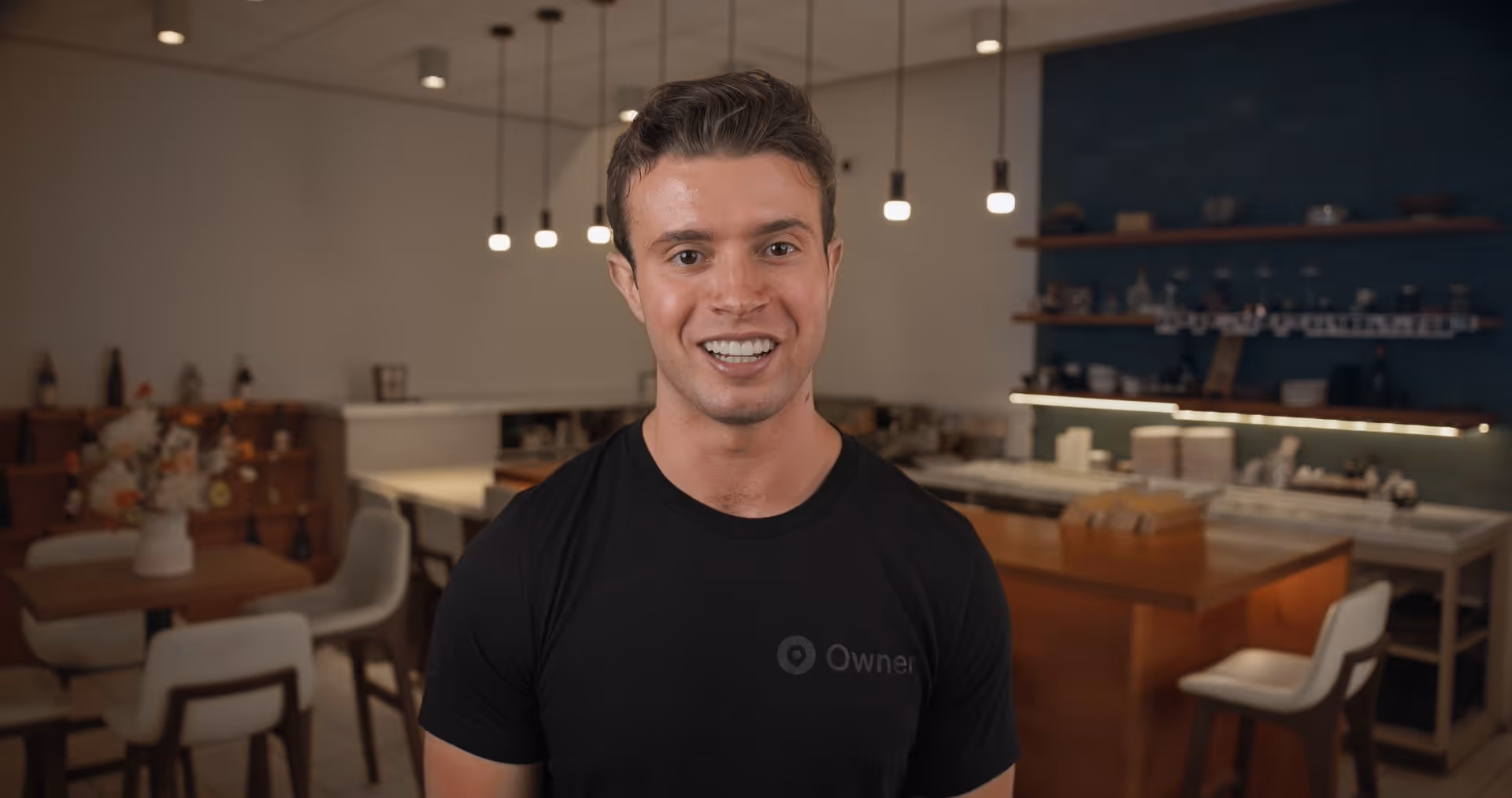 Smiling young man in a black t-shirt standing in a warmly lit modern kitchen with hanging lights.