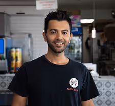 Smiling man wearing a black t-shirt standing in a casual restaurant kitchen.