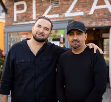 Two men standing close together in front of a brick building with a pizza sign, one with his arm around the other.