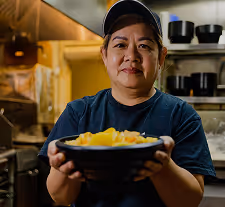 Woman in a kitchen holding a bowl of chopped yellow fruit or vegetables.