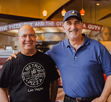 Two men smiling in a pizza restaurant kitchen, one wearing a Metro Pizza Las Vegas shirt and the other in a blue polo and black cap.