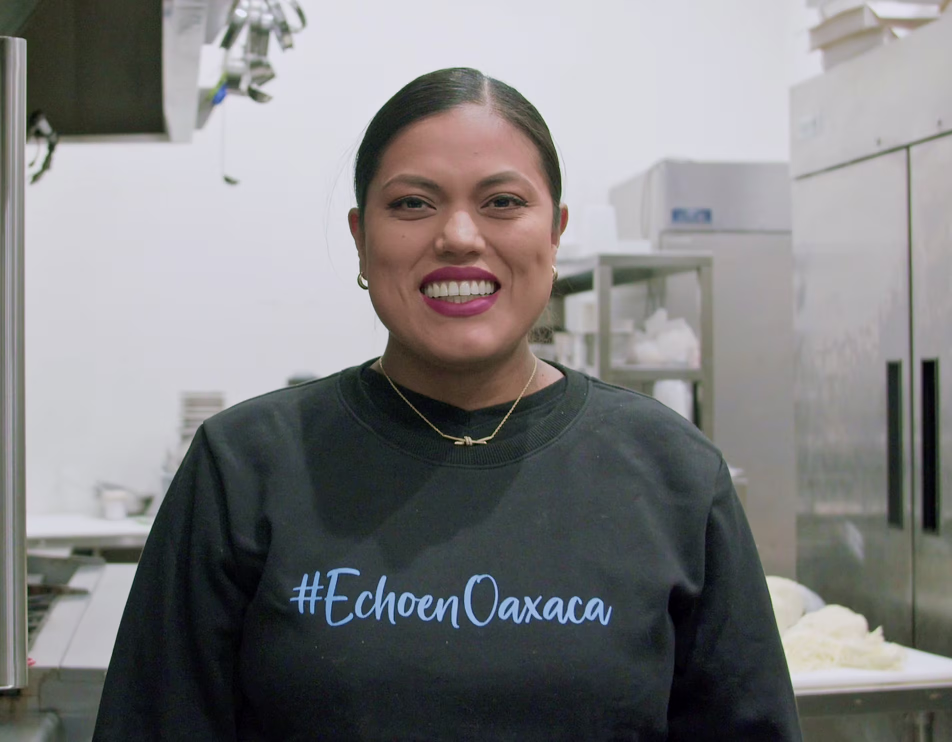 Smiling woman wearing a black sweatshirt with #EchoenOaxaca text, standing in a kitchen.