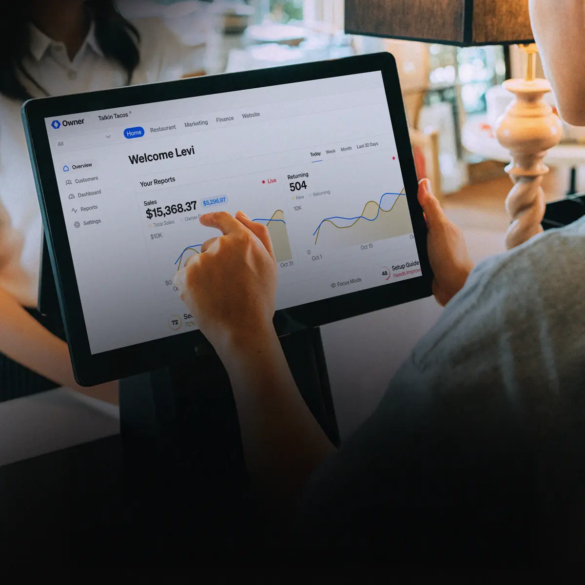 Person using a touchscreen tablet showing a dashboard with sales report of $15,368.37 and returning customers at 504.