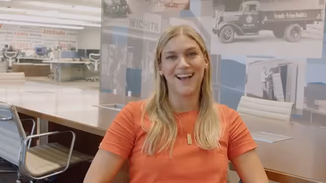 Smiling woman with long blonde hair wearing an orange shirt sitting in a modern office with white chairs and a glass wall.