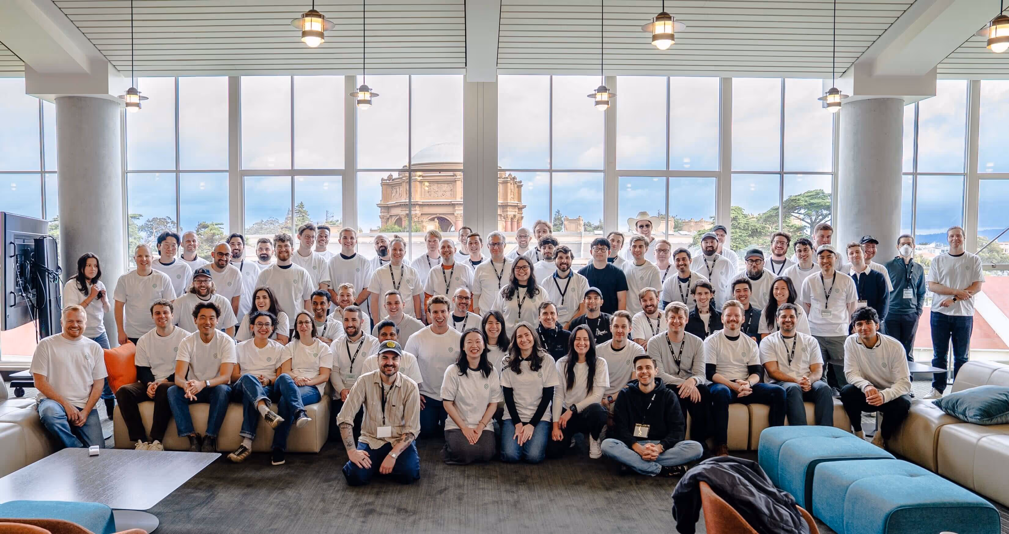 Large group of people posing indoors in front of floor-to-ceiling windows with a historic building visible outside.