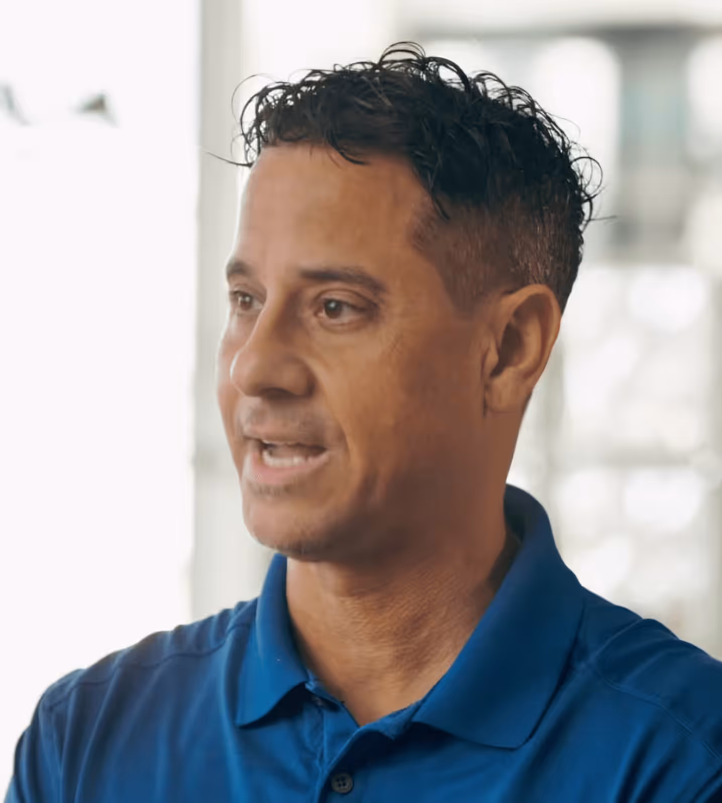 Man with short curly dark hair wearing a blue collared shirt speaking indoors with blurred windows in the background.