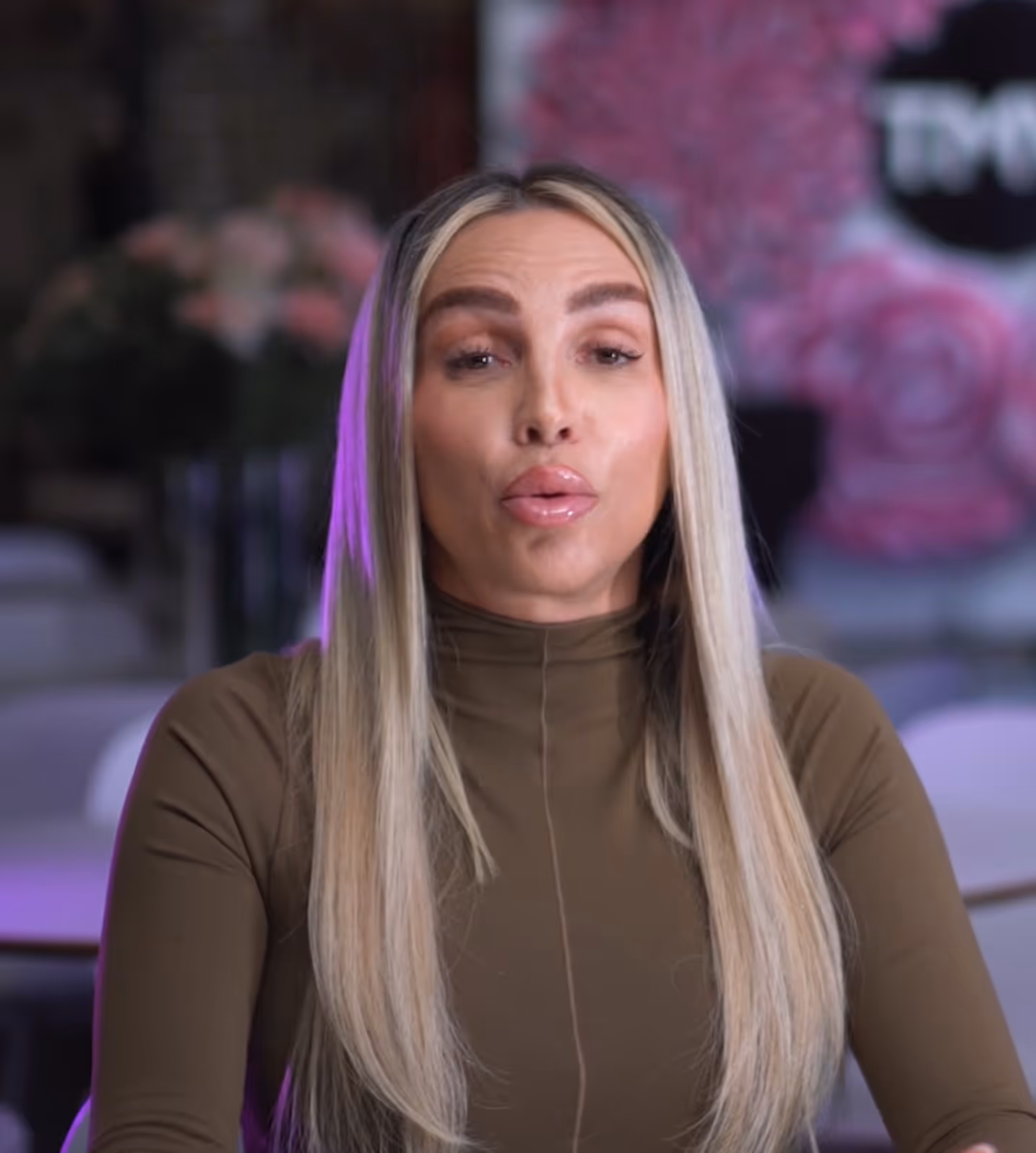 Woman with long blonde hair wearing a brown high-neck top, speaking in an indoor setting with blurred floral background and purple lighting.