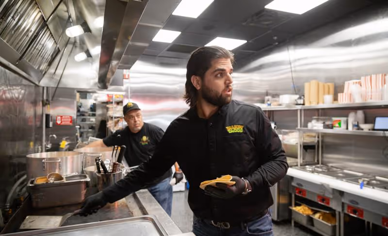 Two men in black uniforms working in a commercial kitchen, one holding a spatula and burger buns, the other near a stove.