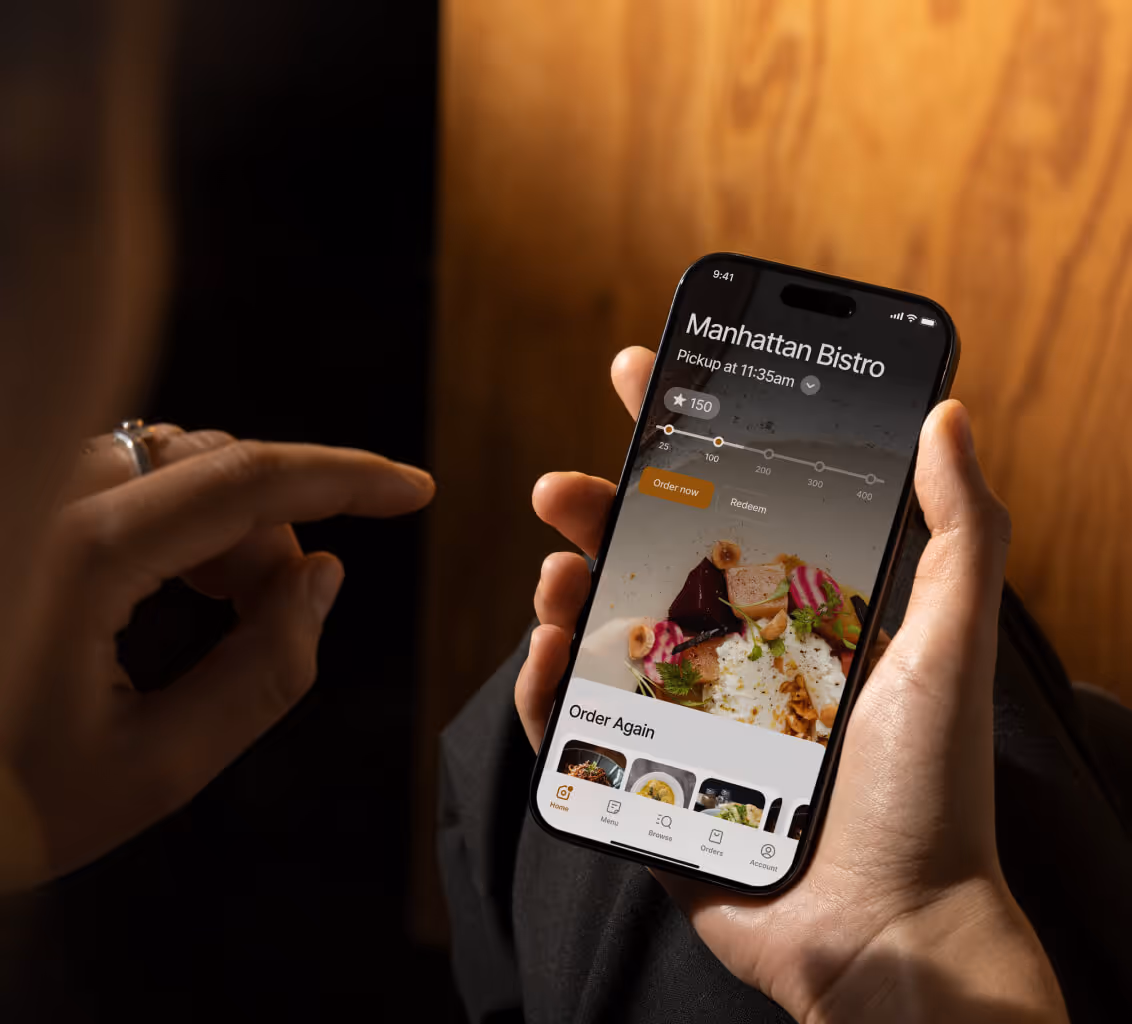 Person holding smartphone displaying a Manhattan Bistro app screen with food ordering options and pickup time.