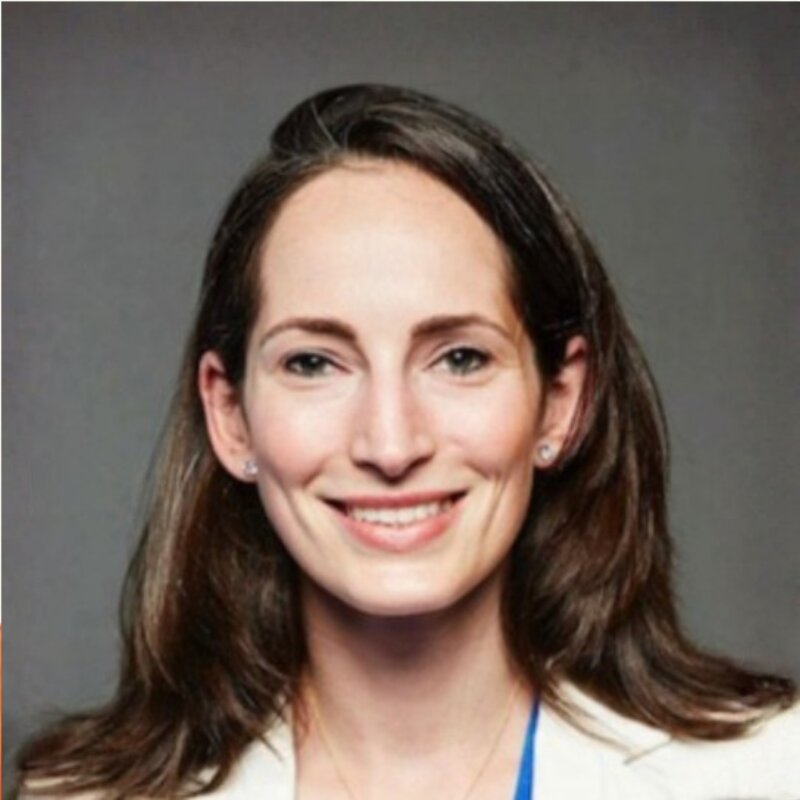 Smiling woman with long brown hair wearing a white blazer and blue top against a gray background.