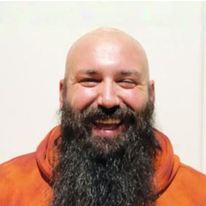 Smiling bald man with a long, thick beard wearing an orange hoodie against a plain light background.