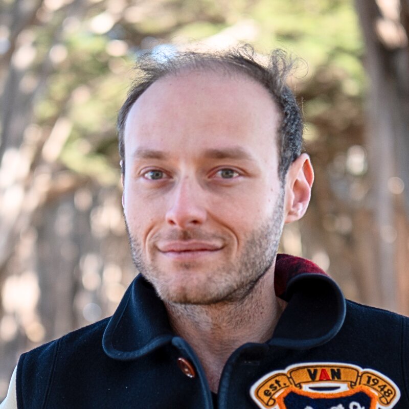 Portrait of a man with short, thinning hair and light facial hair wearing a dark coat outdoors with blurred trees in the background.