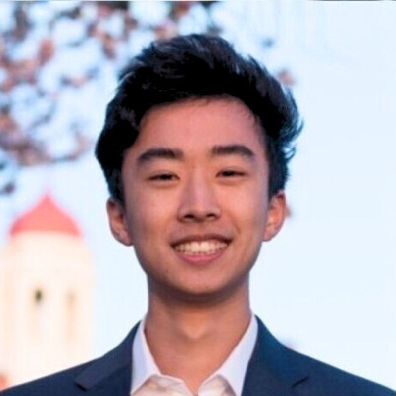 Young man with dark hair wearing a suit jacket and white shirt, smiling outdoors with a blurred building and tree in the background.