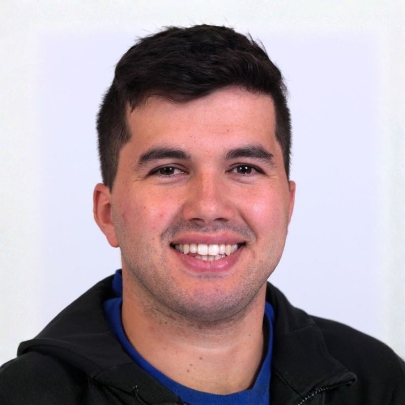 Smiling young man with short dark hair wearing a black jacket and blue shirt against a plain white background.