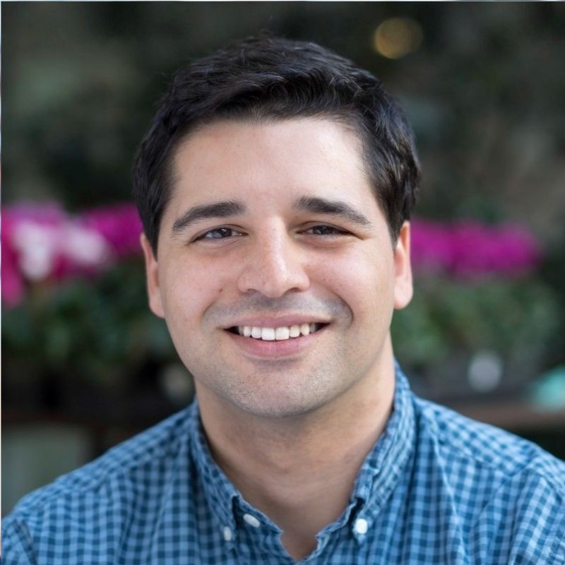 Smiling man with dark hair wearing a blue checkered shirt, with blurred plants and pink flowers in the background.