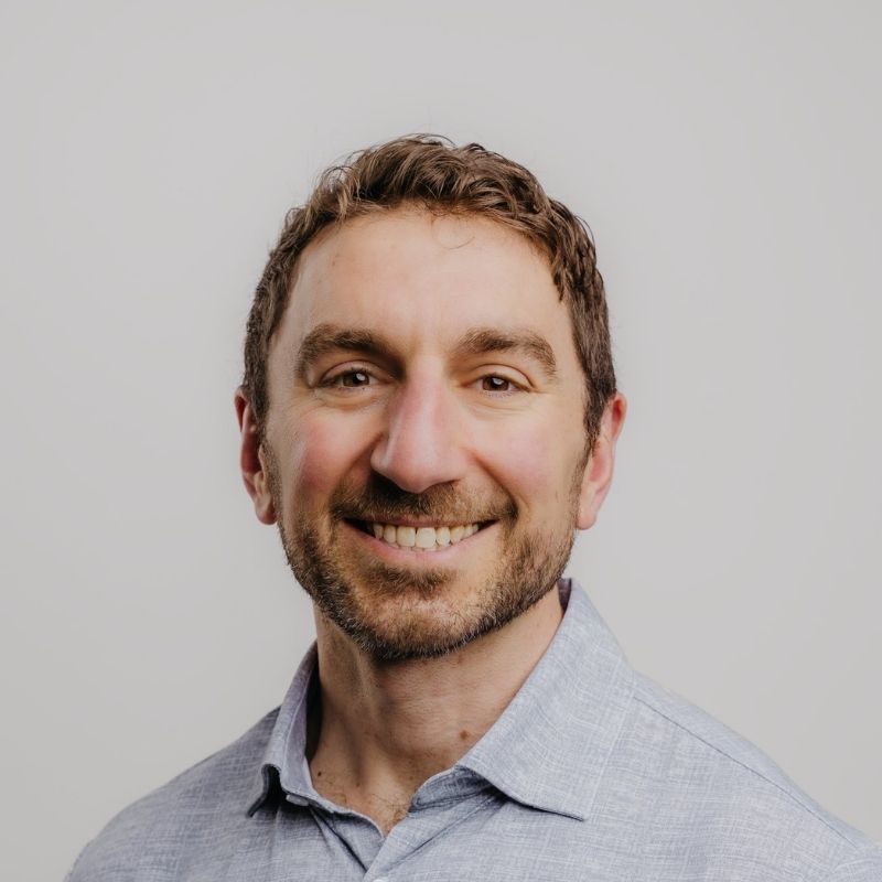 Smiling man with short brown hair, beard, and light blue collared shirt against a light gray background.
