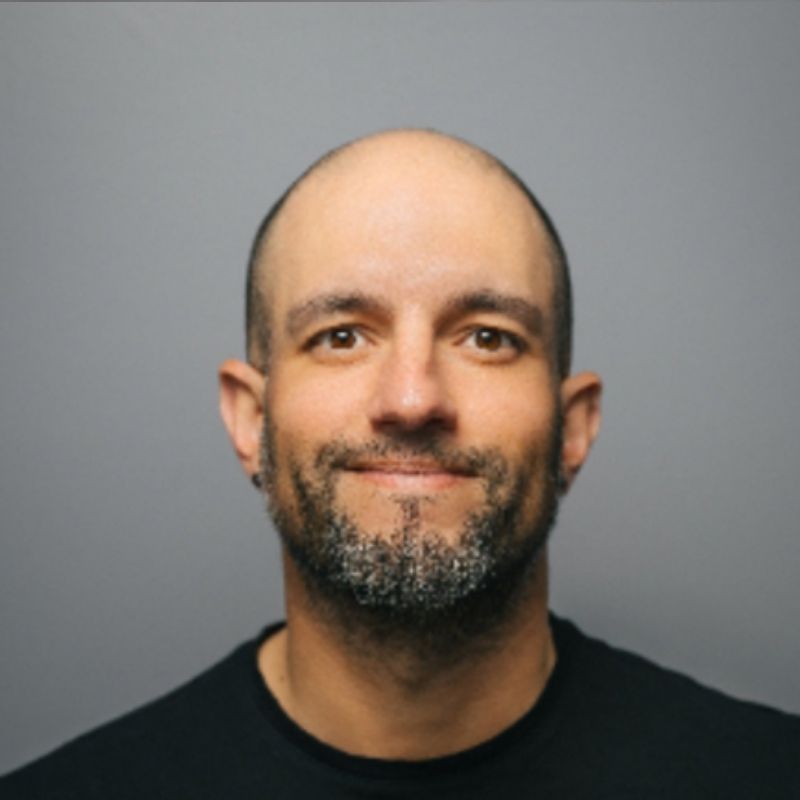 Portrait of a smiling bald man with a short graying beard wearing a black shirt against a gray background.