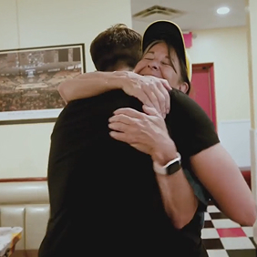 Two men wearing black t-shirts standing together in a kitchen setting, smiling at the camera.
