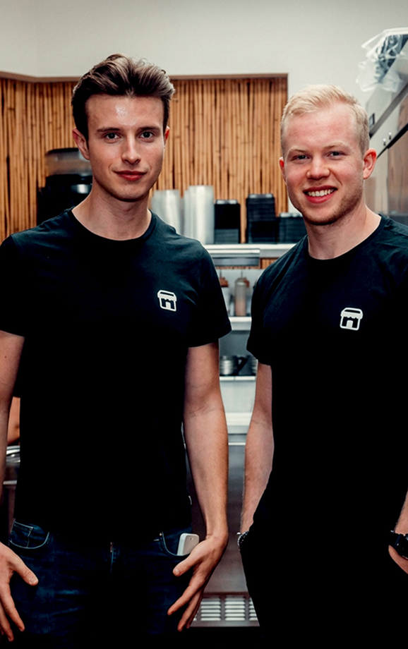 Smiling young man in a black shirt serving noodles with tongs into a white bowl in a kitchen.