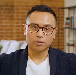 Man with short black hair and glasses wearing a white shirt and dark cardigan speaking indoors with brick wall and bookshelf in background.