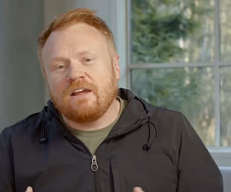 Man with red hair and beard speaking indoors near a window with green trees outside.