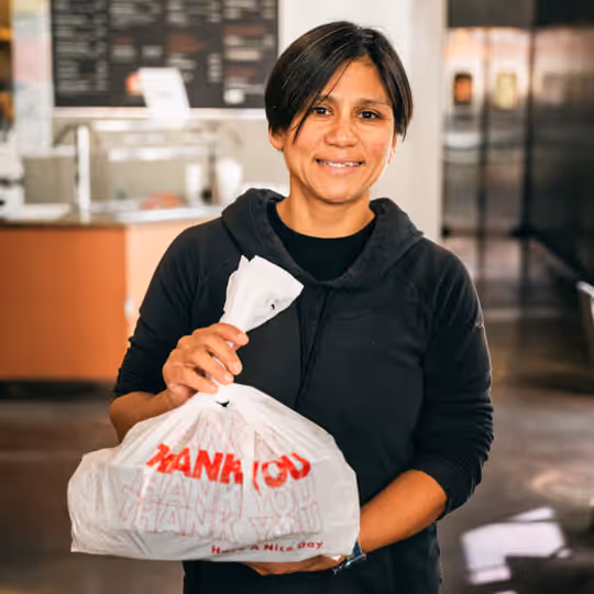 Smiling woman in a black hoodie holding a white plastic bag with red text in a cafe setting.