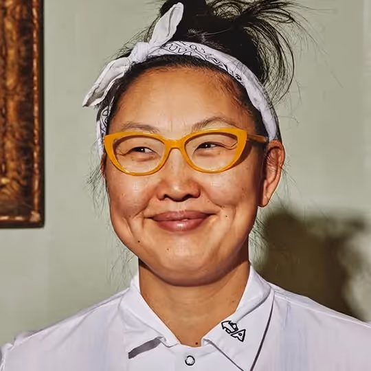 Smiling woman with yellow glasses and a white bandana tied around her head, wearing a white shirt.