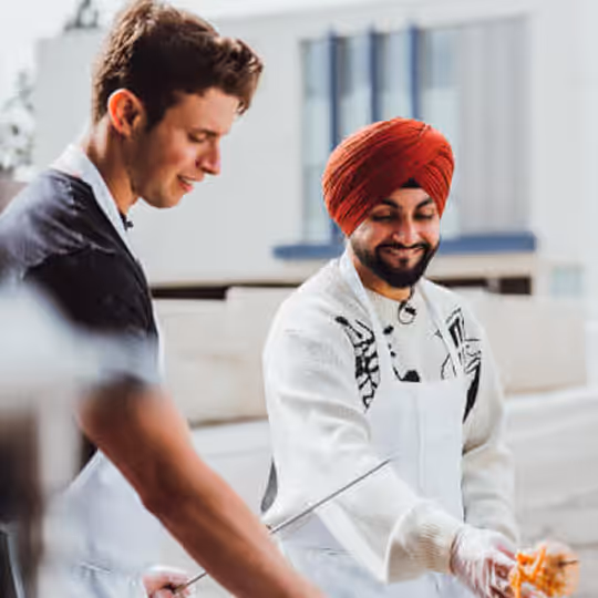 Two men smiling and grilling food outdoors, one wearing a red turban and white apron.