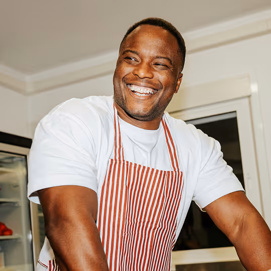 Smiling man wearing a red and white striped apron and white t-shirt in a kitchen.