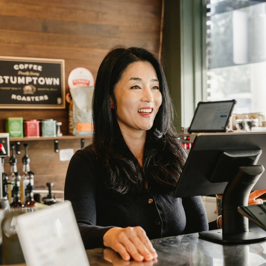 Smiling barista with long black hair standing behind the counter in a coffee shop with Stumptown Roasters sign in the background.