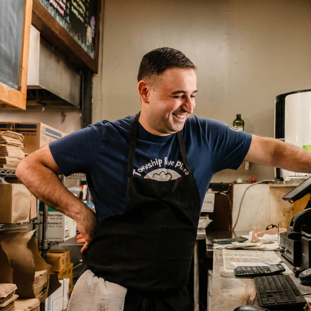Smiling man wearing a blue shirt and black apron standing behind a counter in a kitchen or food service area.