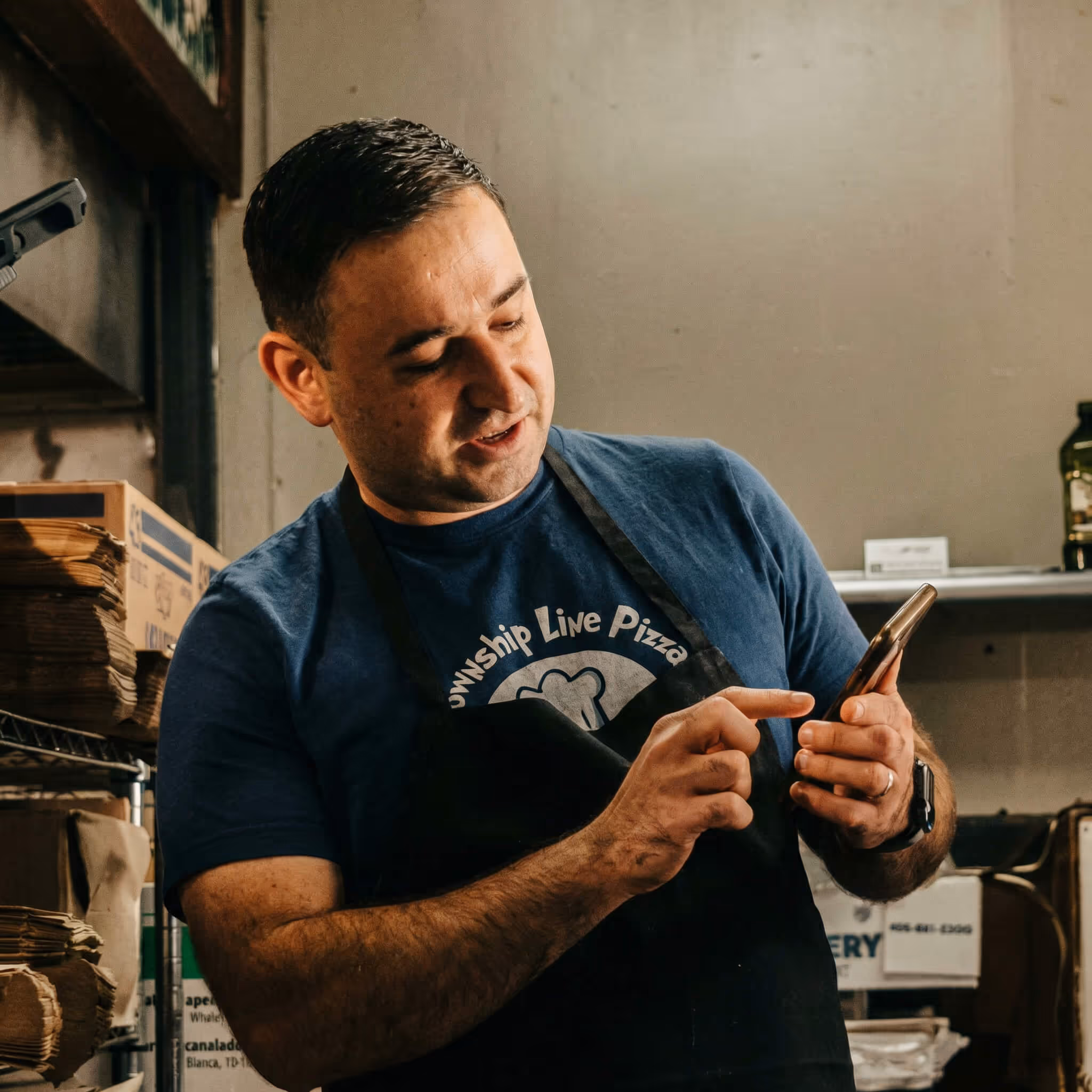 Man in a pizza shop apron using a smartphone inside a storage room with boxes and shelves.