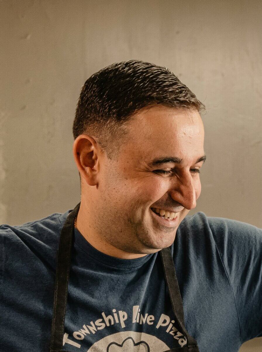Smiling man with short dark hair wearing a dark blue Township Line Pizza t-shirt and apron.