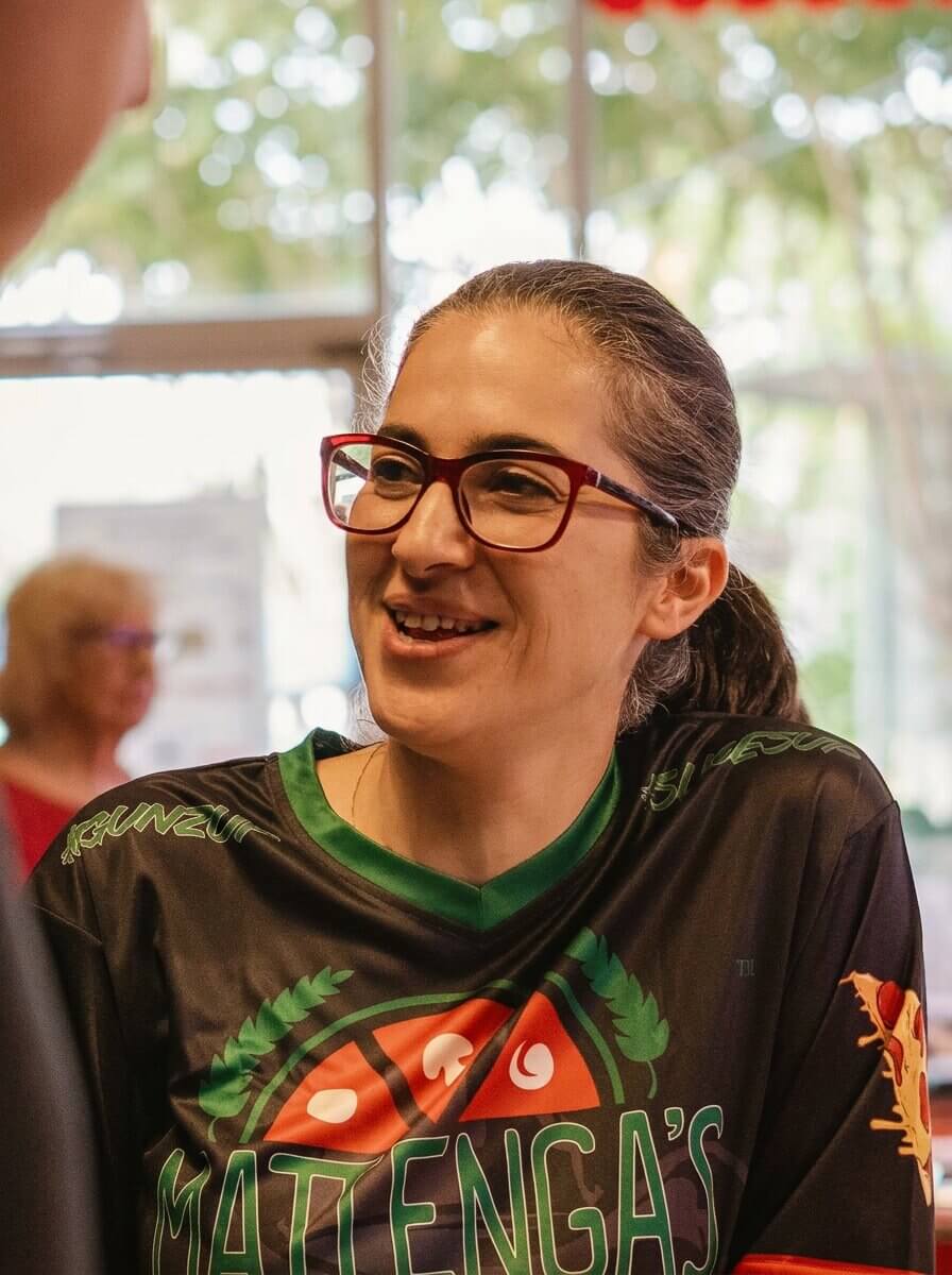 Woman with dark hair tied back and red glasses smiling and wearing a black shirt with green trim and a colorful Mattenga's logo.