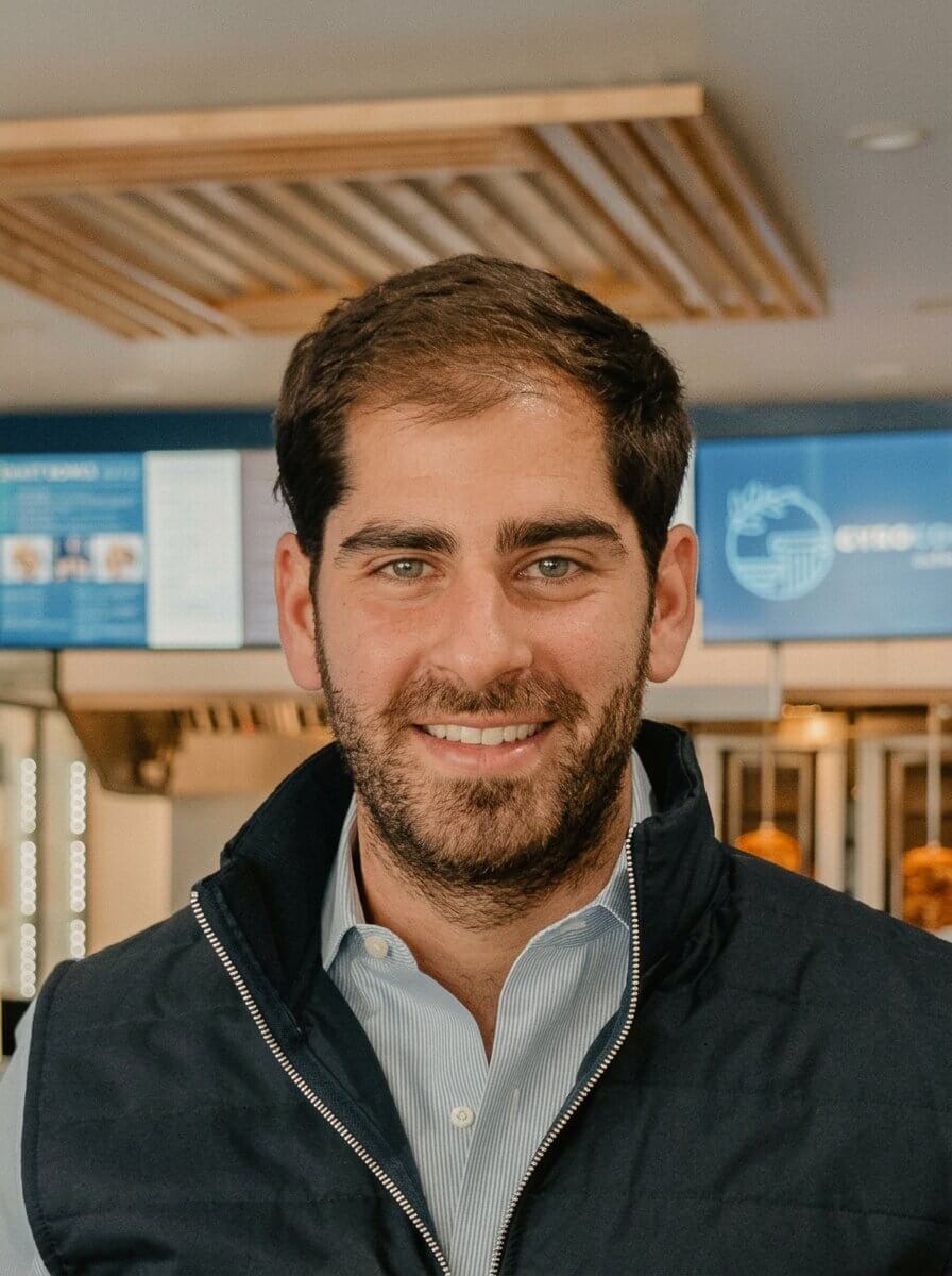 Smiling man with short dark hair and beard wearing a navy jacket and light blue dress shirt indoors.