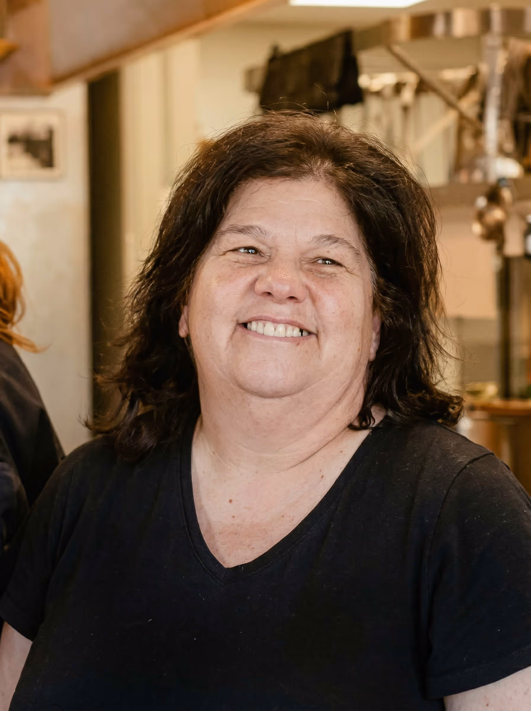 Smiling woman with dark hair wearing a black V-neck shirt in a warm indoor setting.